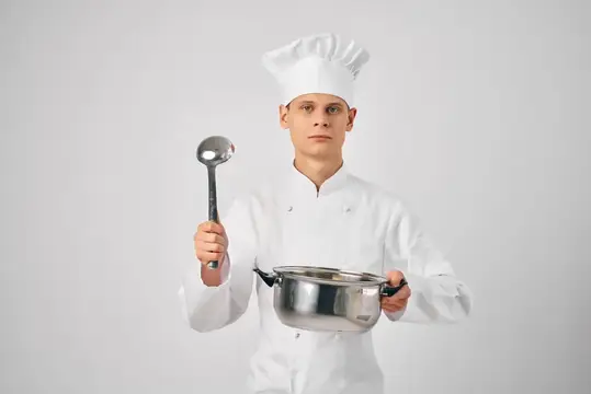 Мужчина в поварской форме держит в руках сковороду и половник, готовя еду