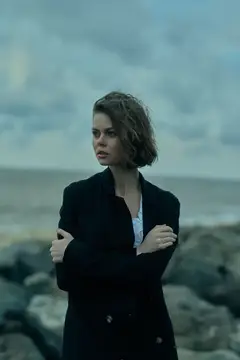 Уличная сцена с женщиной в пальто на берегу, развевающиеся на ветру волосы и виднеющийся вдалеке океан, вызывающие чувства созерцания, стиля и стойкости