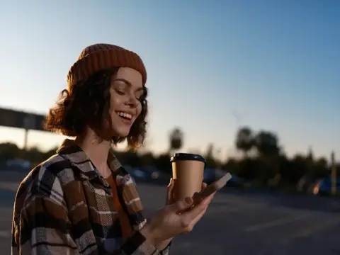 Улыбающаяся девушка в шапке с кофе смотрит в телефон на закате