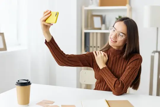 Молодая женщина в очках делает селфи на жёлтый смартфон за столом