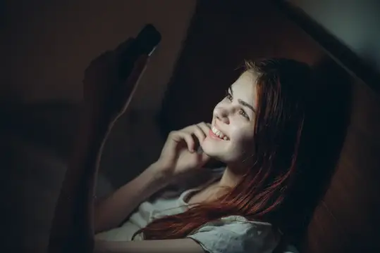 Улыбающаяся девушка лежит в темноте и смотрит в смартфон