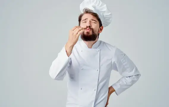 Бородатый повар в белом колпаке пробует блюдо и жестикулирует