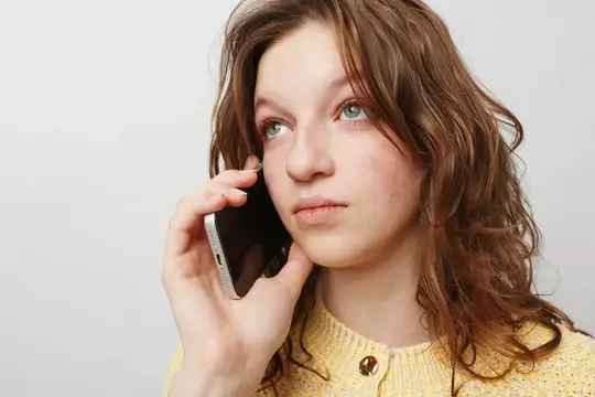 Кудрявая женщина в жёлтом вязаном свитере прижимает смартфон к уху, глядя в сторону с задумчивым выражением лица