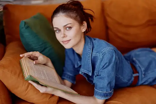 Молодая женщина в джинсовой рубашке лежит на оранжевом диване с книгой