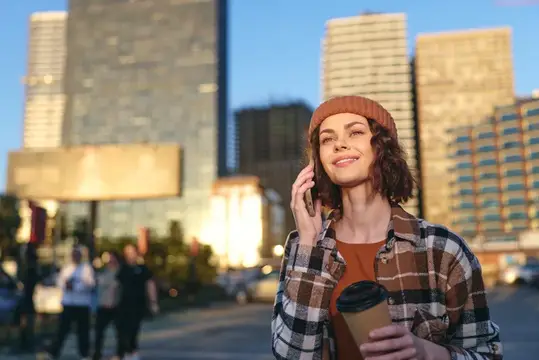 Улыбающаяся девушка в шапке говорит по телефону на фоне вечернего города