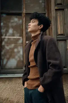 Задумчивый темноволосый парень в пальто смотрит вверх у деревянного дома