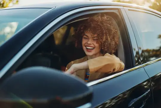 Кудрявая девушка весело смеётся, выглядывая из окна автомобиля