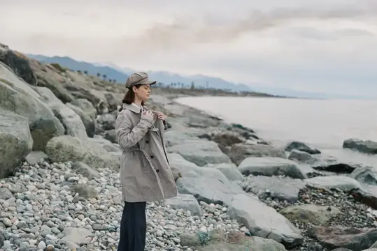 Женщина, одетая в клетчатое пальто и шляпу, стоит на каменистом пляже у спокойного моря