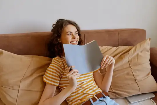 Улыбающаяся темноволосая девушка с блокнотом лежит на диване