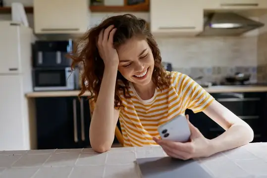 Веселая молодая женщина сидит за кухонной стойкой дома, листает смартфон и делится легким, беззаботным настроением
