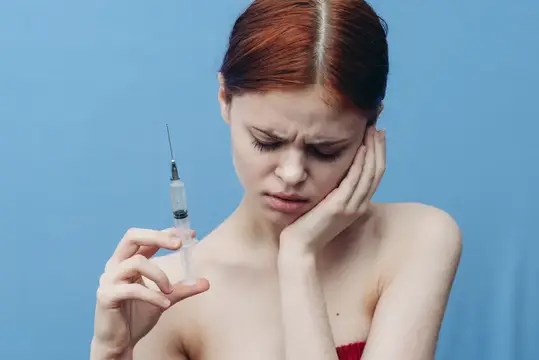 Женщина делает инъекцию, её плечи обнажены, на лице испуганное выражение, на синем фоне