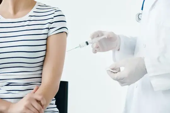 Врач в перчатках делает укол в плечо пациентке в полосатой футболке