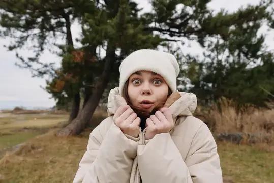 Девушка в белой шапке и пуховике забавно удивляется на природе