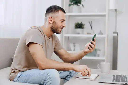 Улыбающийся мужчина смотрит в смартфон, сидя на диване дома