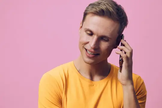 Улыбающийся блондин в жёлтой футболке говорит по телефону на розовом фоне