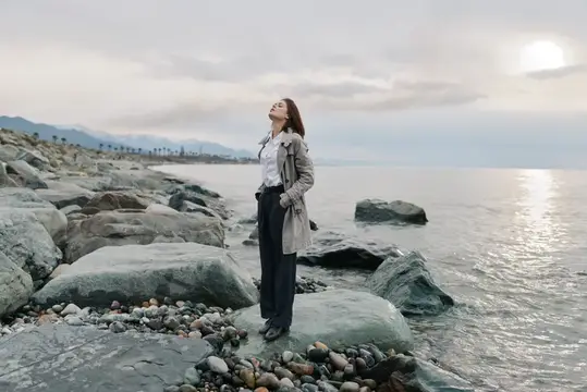 Молодая женщина в плаще стоит на камнях у моря с закрытыми глазами