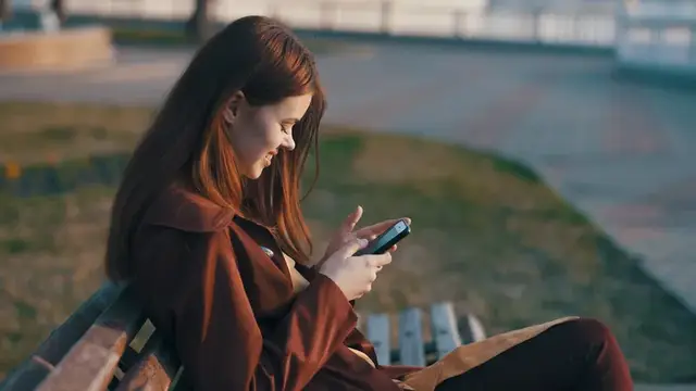 Молодая девушка с длинными волосами улыбается, глядя в смартфон на скамейке