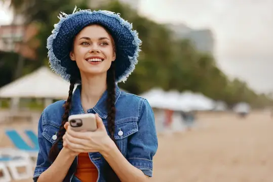 Улыбающаяся девушка в синей шляпе с телефоном на пляже