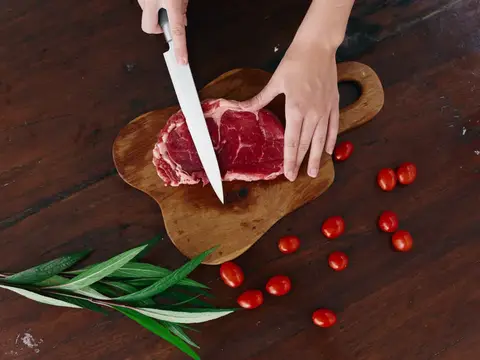 Женщина с ножом в руке нарезает свежий стейк из мяса для жарки на кухне, рядом соль, перец и другие специи на столе, красные помидоры черри и зелень, приготовление ужина