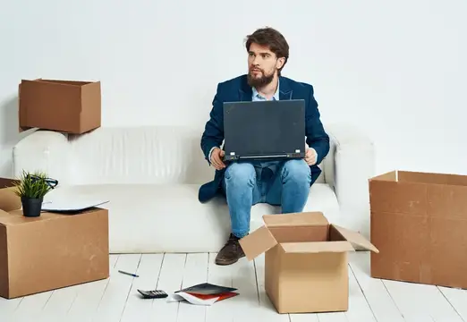 Мужчина сидит на диване перед ноутбуком, работая с коробками вещей, распаковывая новый офис