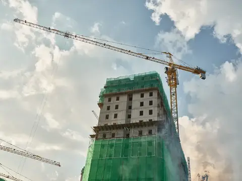Высокое здание в процессе строительства, окутанное зелёной сеткой, с башенным краном на заднем плане на фоне яркого неба с редкими облаками