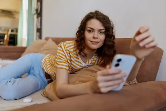 Кудрявая девушка лежит на диване со смартфоном и улыбается