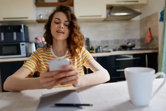 Кудрявая девушка в полосатой футболке смотрит в смартфон на кухне
