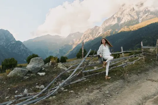 Молодая женщина в походе в горах: одета в белую куртку и брюки, идёт вдоль деревянного забора по каменистой тропе на фоне драматичных облаков и сурового альпийского пейзажа