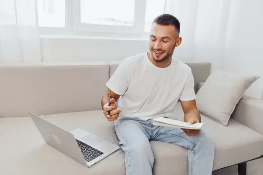Улыбающийся мужчина в расслабленной одежде работает из дома за ноутбуком, держа в руках блокнот