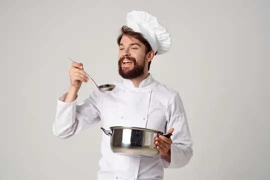 Бородатый повар в колпаке пробует суп из кастрюли