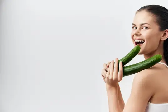 Улыбающаяся девушка с двумя огурцами у лица на белом фоне