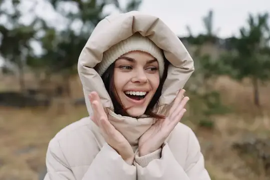Смеющаяся девушка в светлом пуховике и шапке на природе