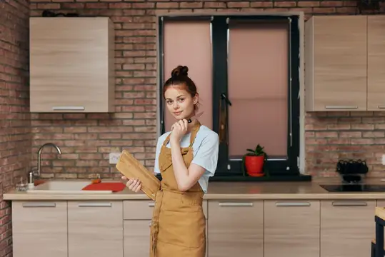 Весёлая женщина в коричневом фартуке режет продукты на разделочной доске на кухне