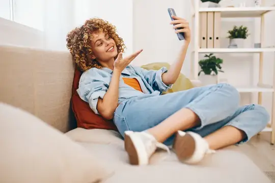 Улыбающаяся женщина держит мобильный телефон на уютном диване в гостиной, наслаждаясь онлайн-играми и общением в счастливом доме