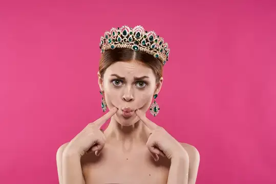 Девушка в короне с зелёными камнями кривляется на розовом фоне