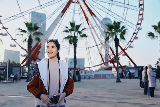 Улыбающаяся азиатская девушка с фотоаппаратом на фоне колеса обозрения