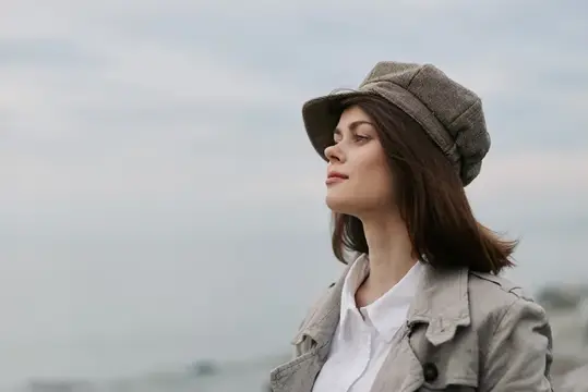 Портрет спокойной женщины в клетчатой кепке и легком пальто, стоящей у океана с облачным небом