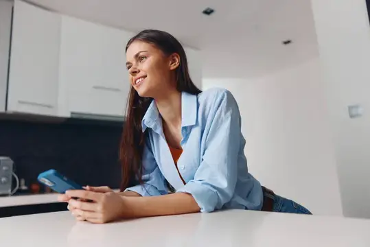 Улыбающаяся девушка со смартфоном облокотилась на стол на светлой кухне