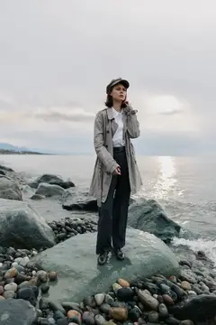 Молодая женщина в плаще и кепке стоит на камне у моря