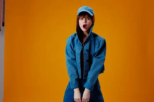 Удивлённая девушка в джинсовой куртке и кепке на оранжевом фоне