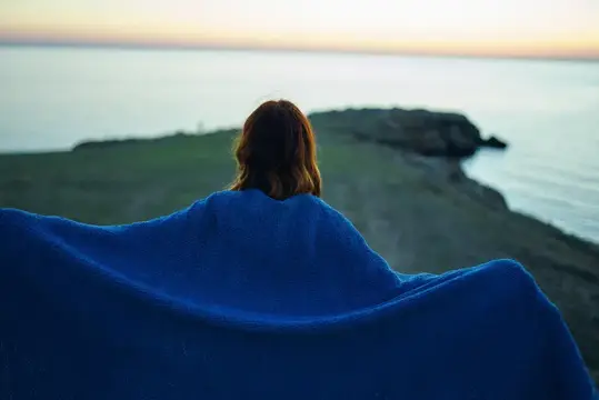 Девушка в синем пледе смотрит на море и скалы
