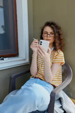 Кудрявая девушка в очках смотрит в смартфон, сидя в кресле у окна