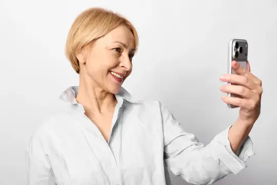 Улыбающаяся блондинка в белой рубашке делает селфи на смартфон