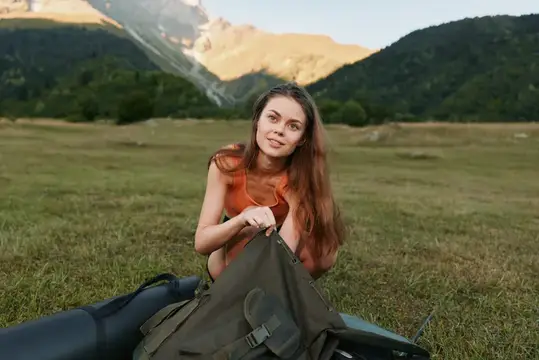 Молодая женщина с рюкзаком сидит на траве на фоне гор