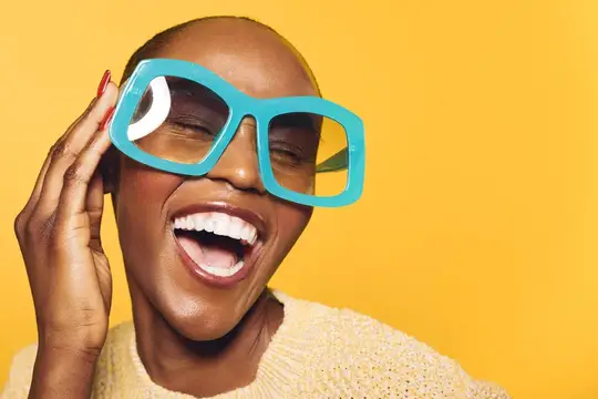 Темнокожая женщина в огромных бирюзовых очках смеётся на жёлтом фоне