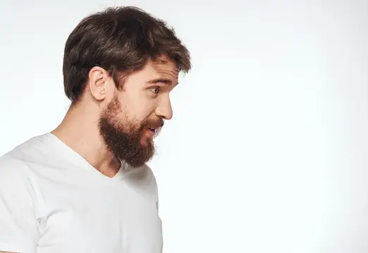 Бородатый мужчина в белой футболке удивлённо смотрит в сторону на светлом фоне