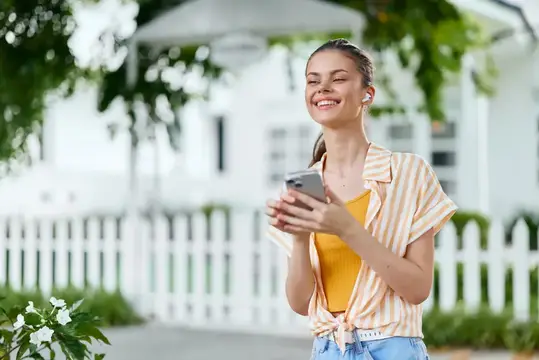 Улыбающаяся девушка в наушниках держит смартфон у белого забора