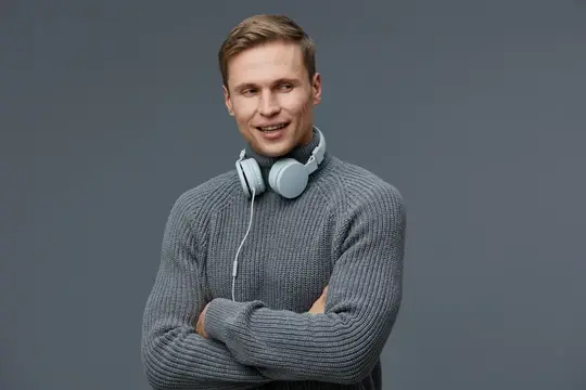 Улыбающийся светловолосый мужчина в сером свитере с наушниками на шее
