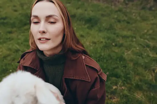 Молодая женщина, одетая в коричневую куртку и тёмно-зелёный свитер, нежно держит белого козлёнка, стоя на зелёной траве, изображая мирную и аутентичную жизненную сцену