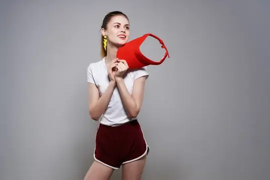 Улыбающаяся девушка в белой футболке держит красную сумку на сером фоне
